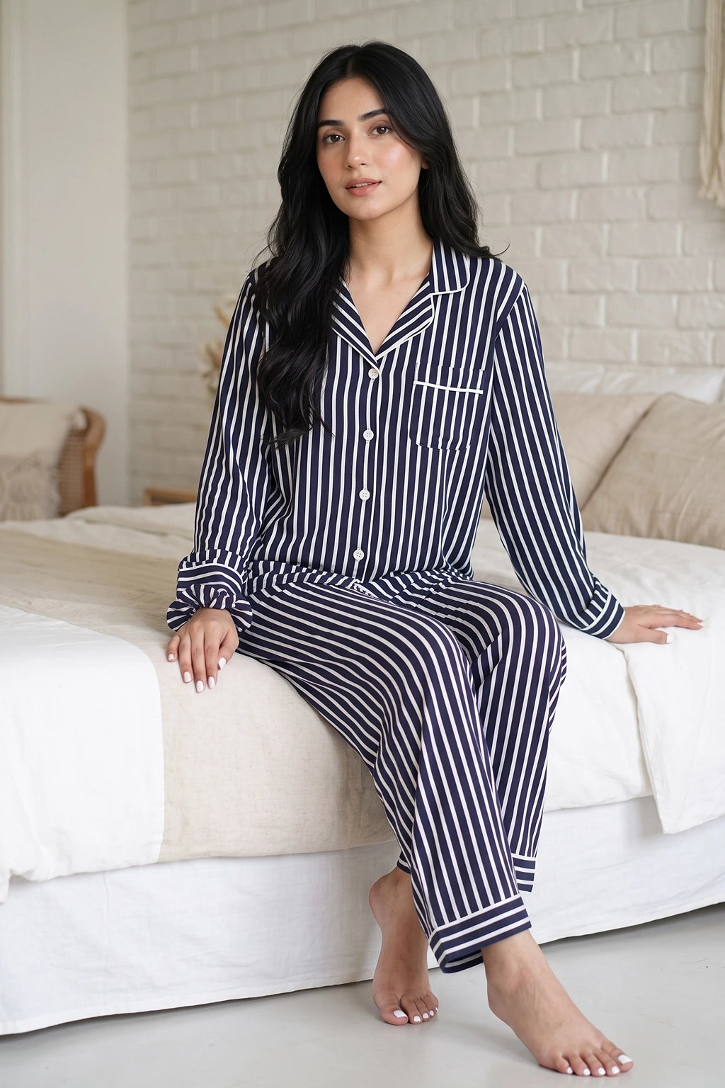 A woman wearing a navy and white vertically striped long-sleeve linen pajama set with a button-up front, sitting on a white bed in a bedroom setting.