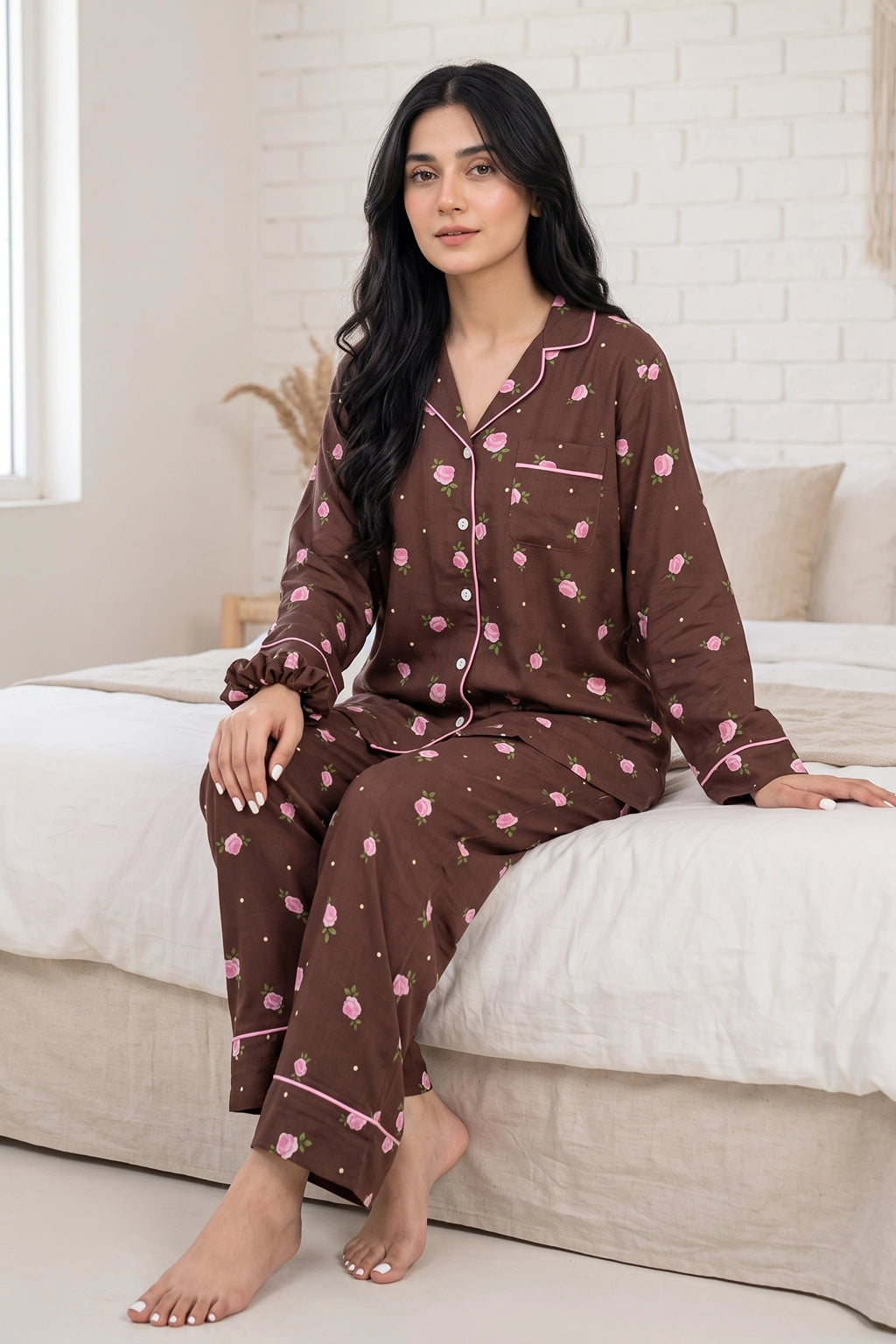 A woman wearing a brown long-sleeve pajama set with a pink floral pattern sits on a bed with white linens in a bright bedroom.
