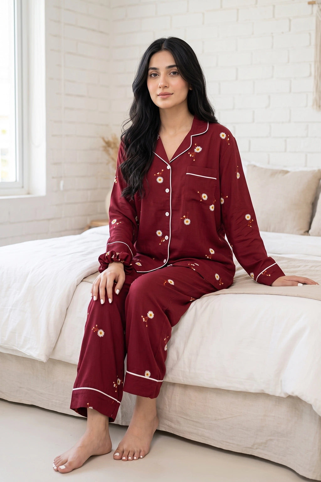 A Pakistani woman wearing a burgundy long-sleeve linen pajama set with white piping and small floral prints, sitting on a bed in a bright bedroom.