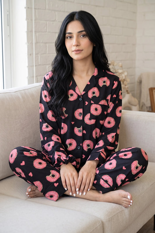 A Pakistani woman sits cross-legged on a beige sofa wearing a black linen printed pajama set with a pink donut and heart pattern.